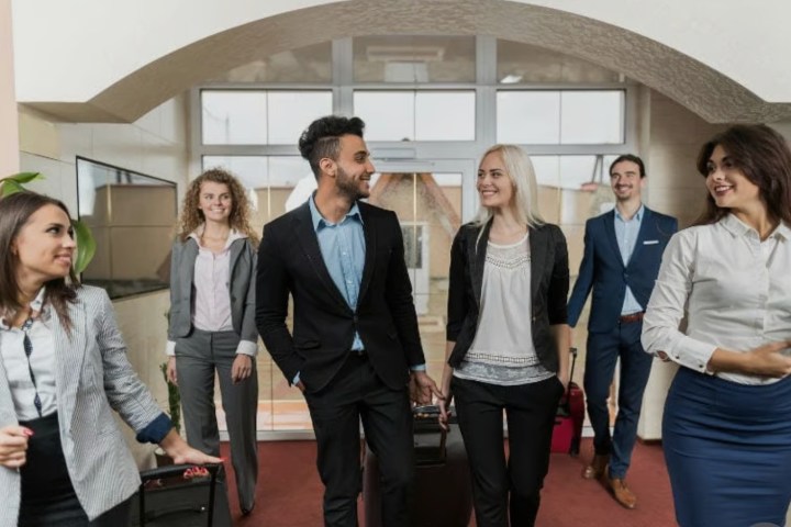 Group of six professionals in business attire walking with luggage indoors.